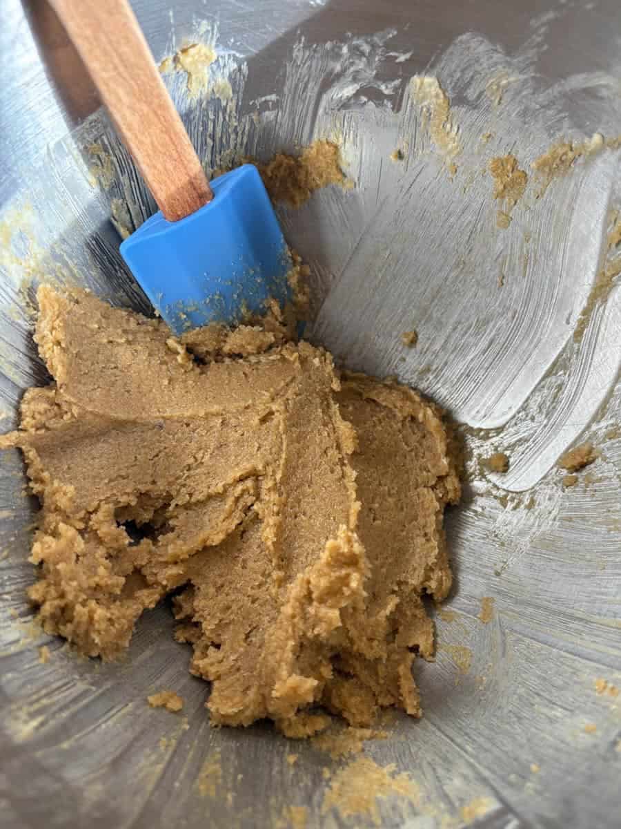 A silver mixing bowl of creamed butter, brown sugar and egg with a blue wooden handled spatula.