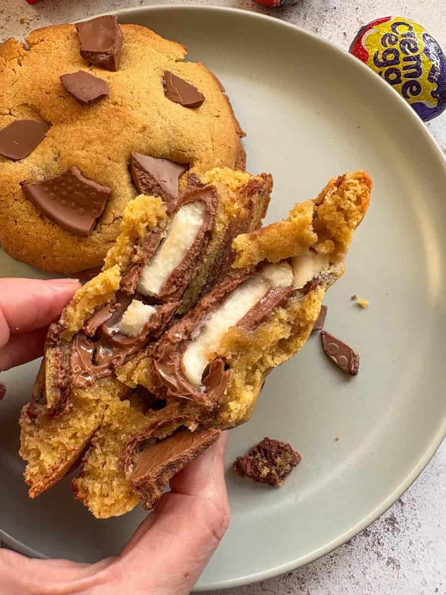 A hand holding a large cookie broken in half to reveal a creme egg middle. Another cookie sits on a grey plate.