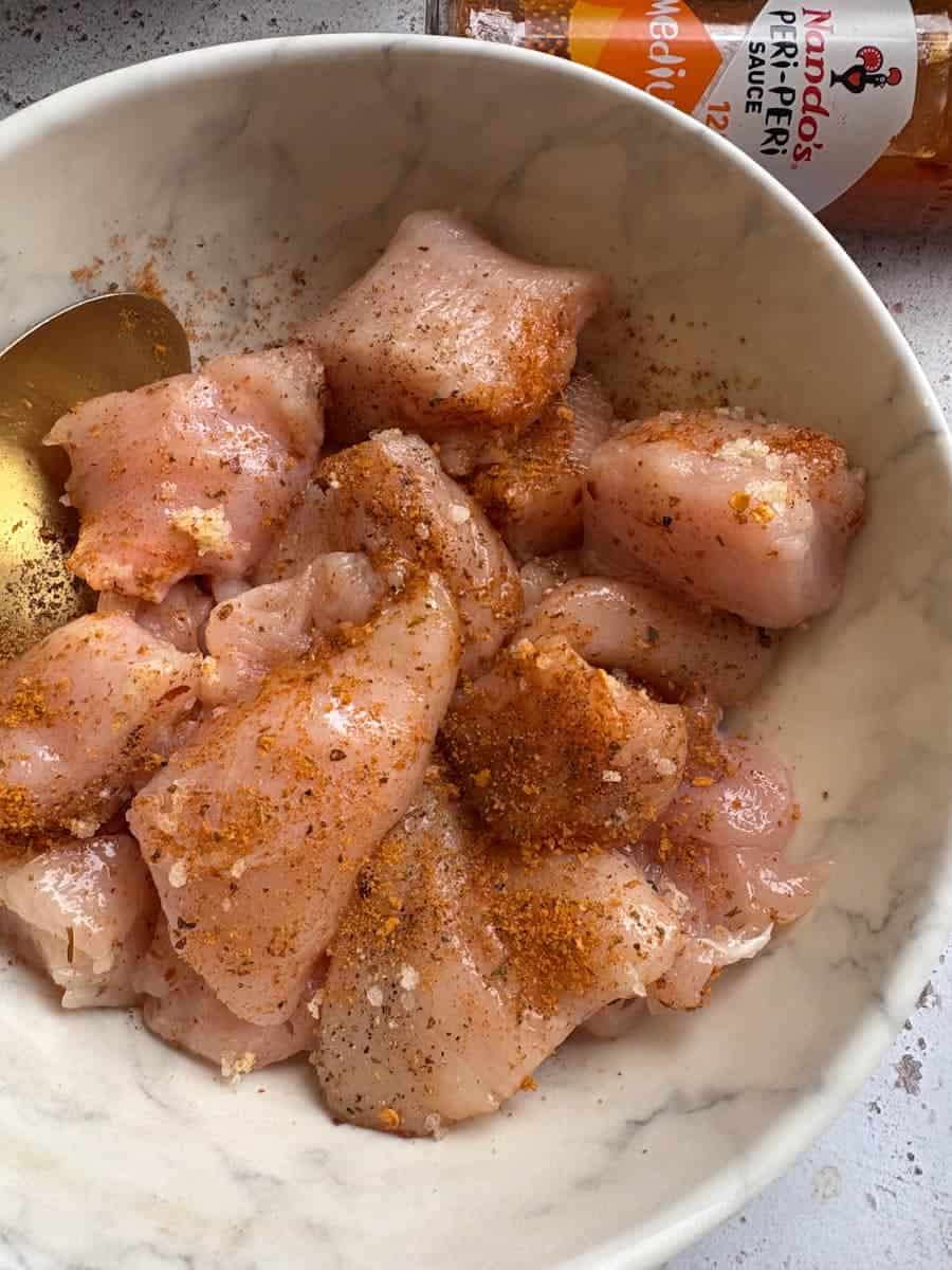 Raw pieces of chicken breast in a white bowl. The chicken has been coated in Peri Peri seasoning.