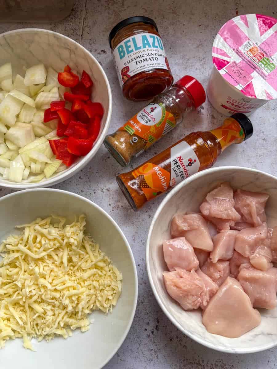 White bowls of diced raw chicken breast, grated mozzarella cheese, diced onion and red peppers, a tub of sour cream, a jar of tomato paste, a bottle of Peri Peri sauce and a jar of Peri Peri seasoning.