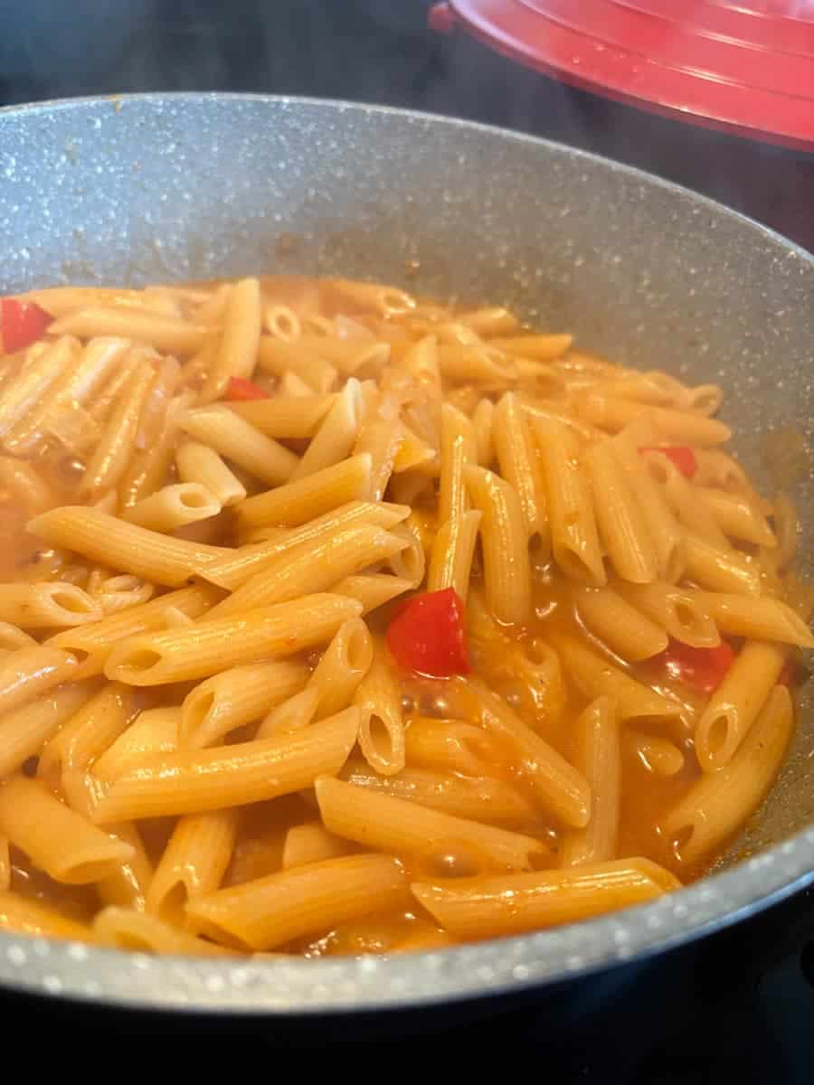 A grey pan of penne pasta with diced red peppers and chicken stock.