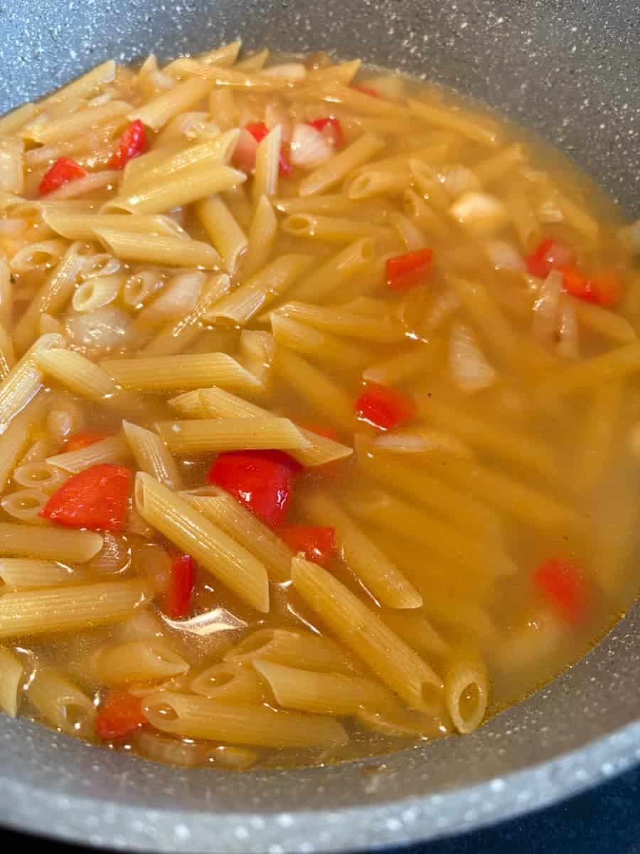 A grey pan of penne pasta with chicken stock, diced red peppers and onion.