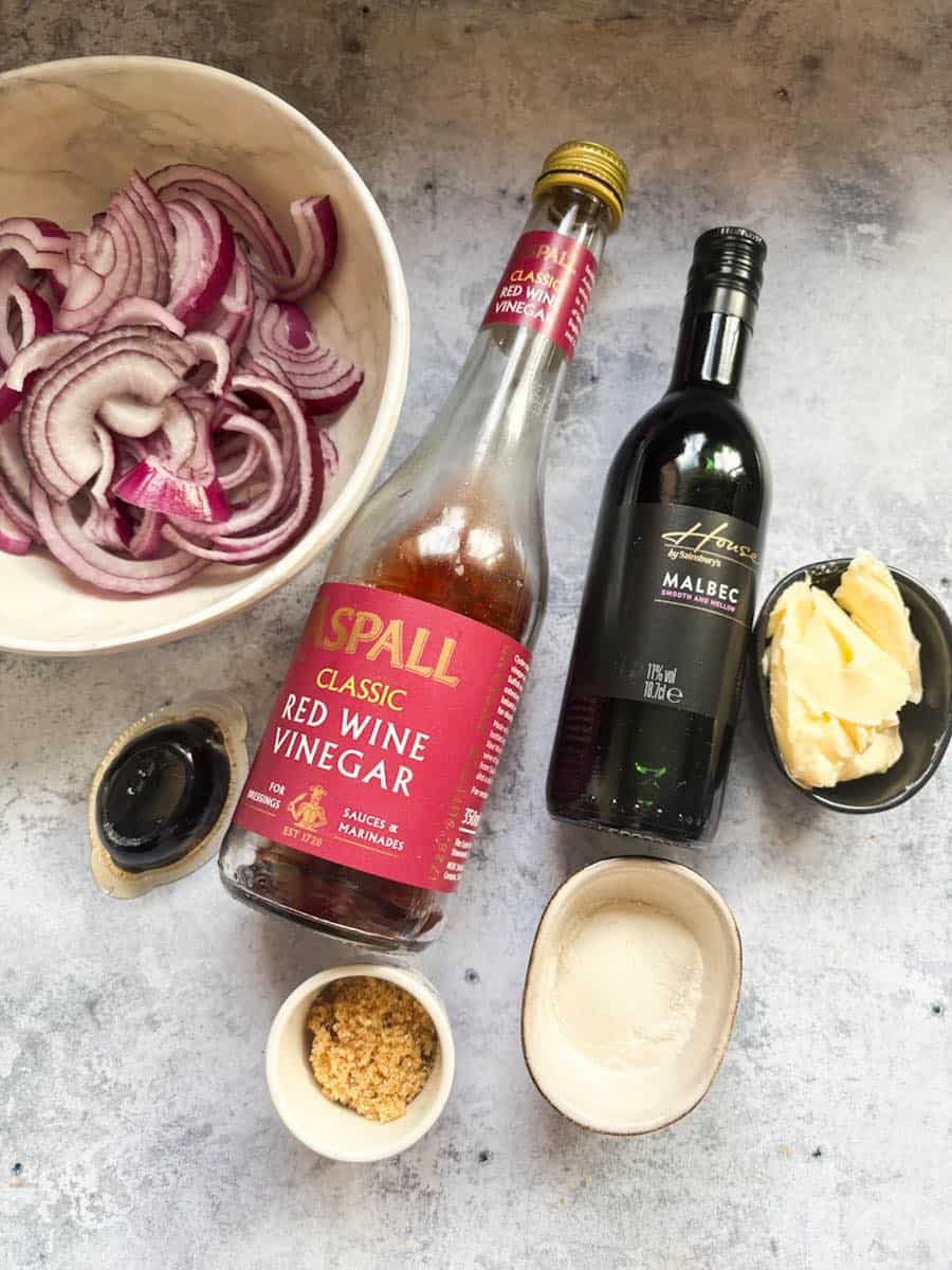 A white bowl of sliced red onions, a bottle of red wine vinegar, a bottle of red wine, a black pot of butter, a beef stock pot, a small pot of brown sugar and a pot of plain flour.