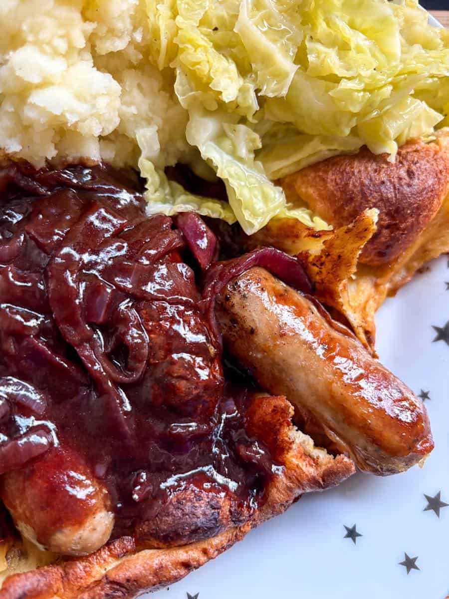Cooked sausages, Yorkshire pudding, red onion gravy, mashed potato and cooked sliced cabbage on a white and gold star plate.