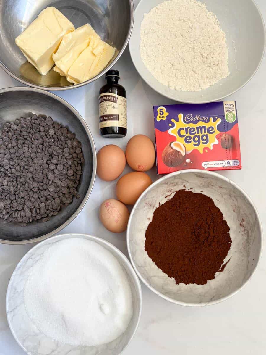 White bowls filled with flour, sugar, cocoa powder, a silver bowl of butter, a bowl of chocolate chips, 4 eggs, a bottle of vanilla extract and a packet of Cadburys Creme Eggs.