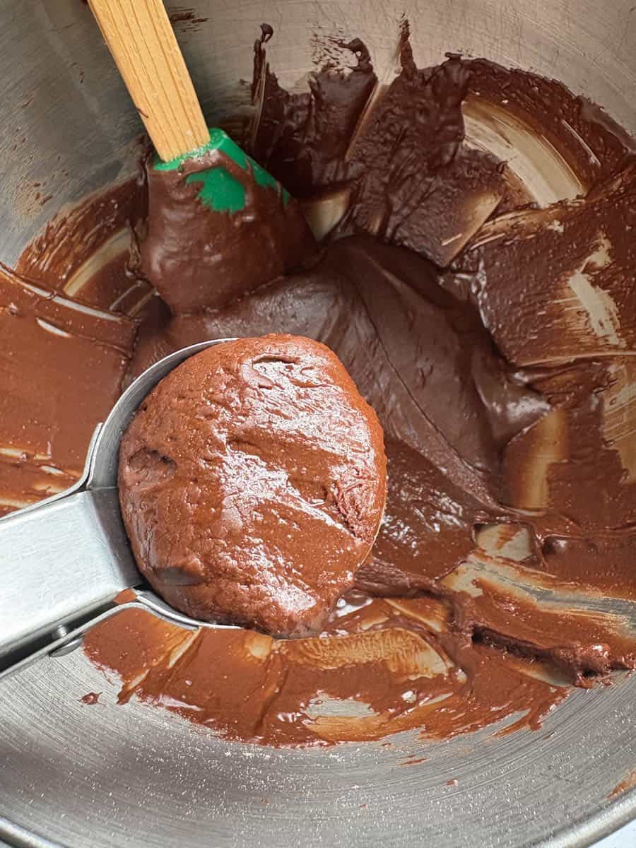 A silver bowl of chocolate brownie cookie batter and an ice cream scoop holding a portion of cookie dough.