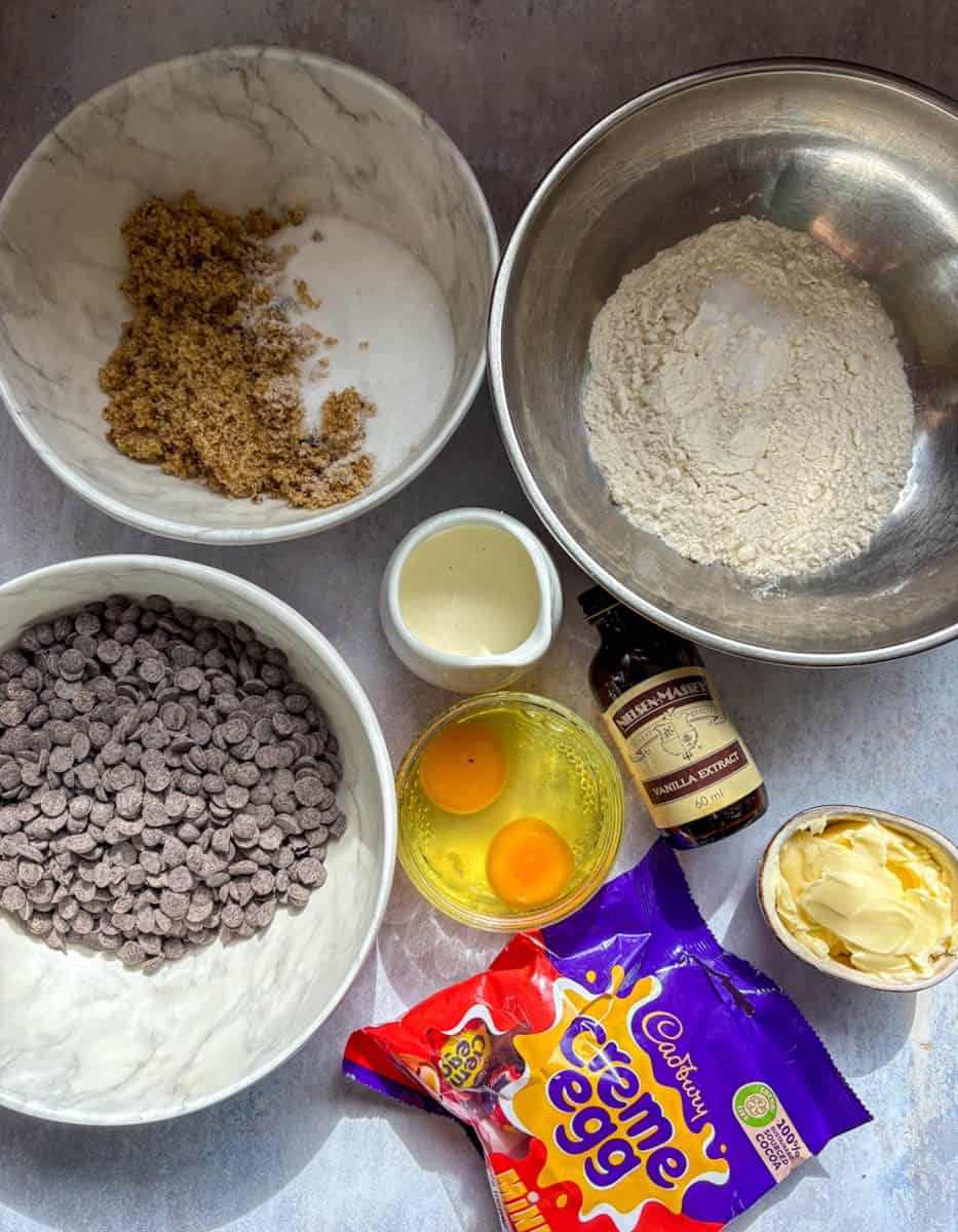 Various ingredients for making chocolate brownie cookies - a silver bowl of flour, a white bowl of brown and white sugar, a white bowl of chocolate chips, two eggs cracked into a glass ramekin, a white jug of cream, a bottle of vanilla extract, a pot of butter and a packet of mini creme eggs.