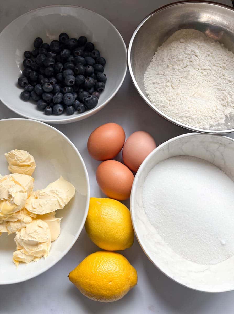 Bowls of white sugar, flour, butter and blueberries, 3 eggs and two lemons on a white surface.