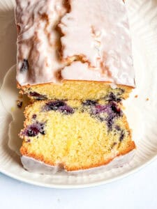 A close up photo of a sliced lemon and blueberry loaf cake covered in white icing on a white plate.