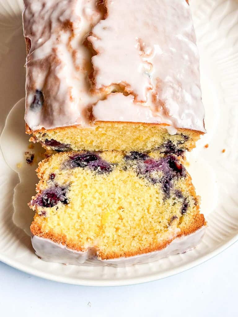 A close up photo of a sliced lemon and blueberry loaf cake covered in white icing on a white plate.