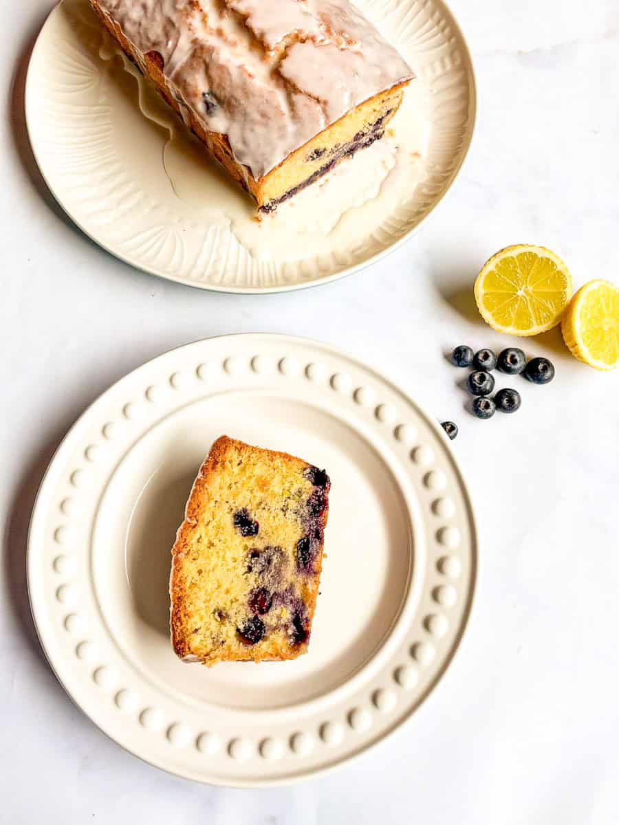 A slice of lemon and blueberry loaf cake on a white plate and a lemon blueberry loaf on a white plate covered in white icing. A few fresh blueberries and a lemon cut in half has been placed beside the plates for decoration.