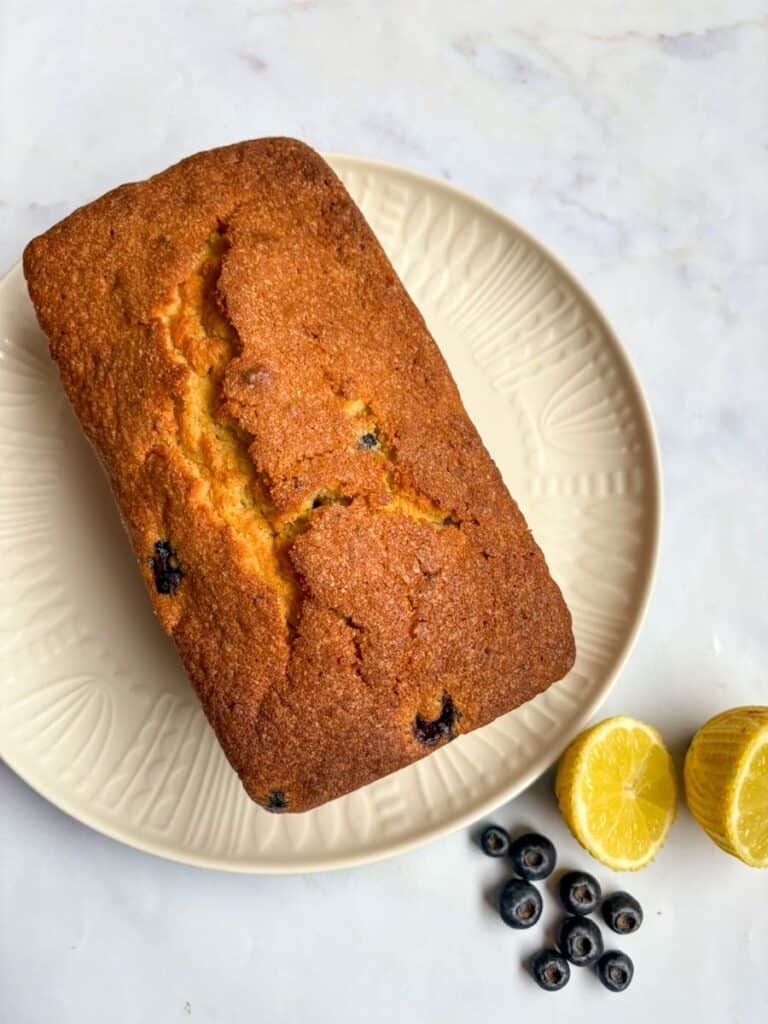 A lemon and blueberry loaf cake on a white plate. Fresh blueberries and a lemon cut in half has been placed beside the plate.