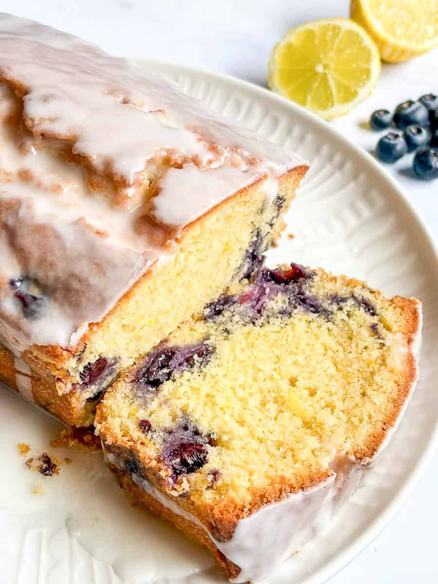A sliced blueberry and lemon loaf cake on a white plate. A lemon cut in half and fresh blueberries can be seen in the background.