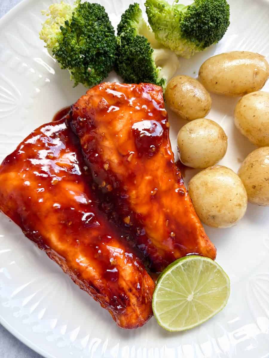 Two salmon fillets covered in sweet chilli marinade on a white plate with new potatoes, steamed broccoli and a slice of lime.