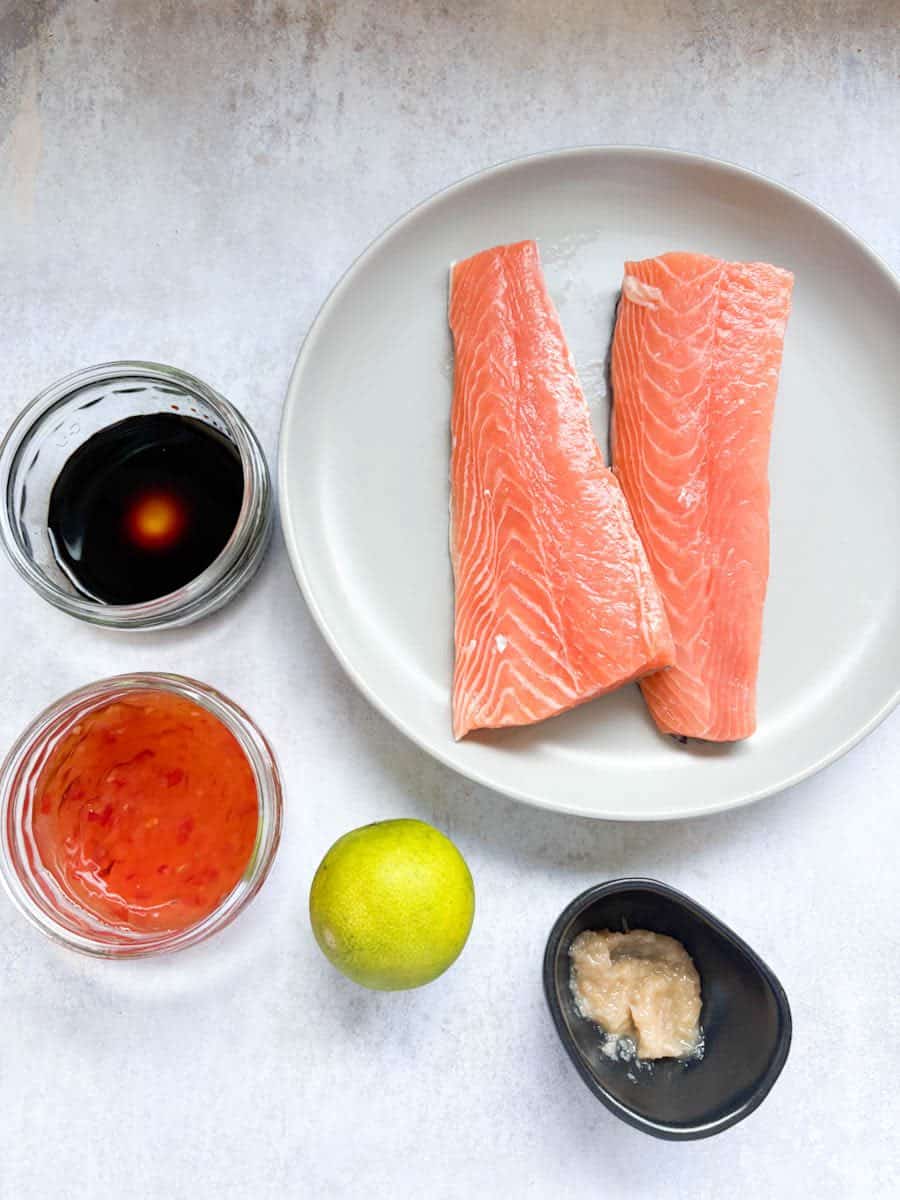 Two salmon fillets on a grey plate, two small glass ramekins filled with soy sauce and sweet chilli sauce, a lime and a black pot of garlic puree.