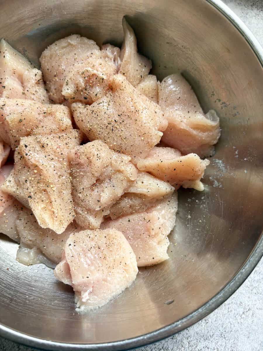 A silver bowl of raw chicken pieces seasoned with salt and pepper.