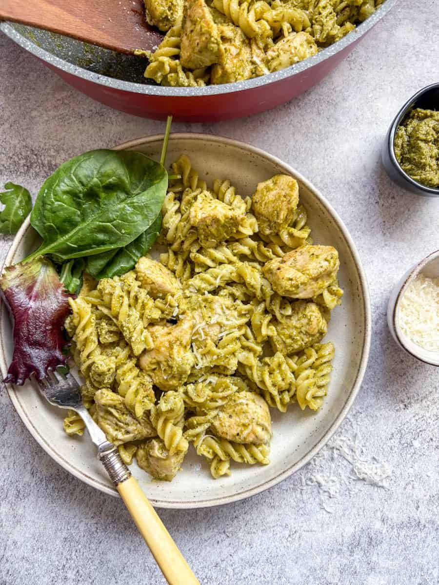 A bowl of chicken pesto pasta with salad leaves and a yellow handled silver fork on a white and grey surface. Small pots of green pesto, grated parmesan cheese and a large pan of pasta can be partially seen.