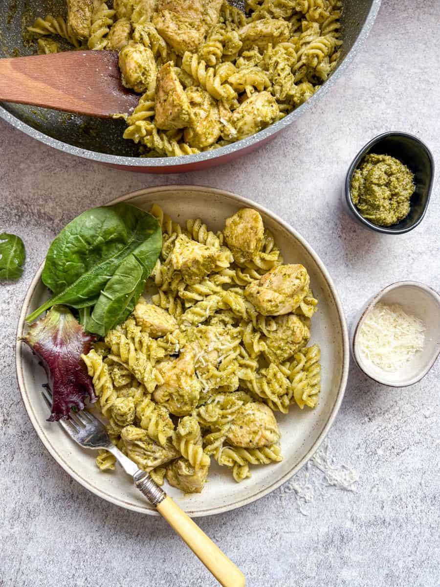 A bowl of chicken pesto pasta with salad leaves on the side and a silver fork with a yellow handle, small pots of green pesto and grated parmesan cheese and a large pan of the pasta with a wooden spoon.