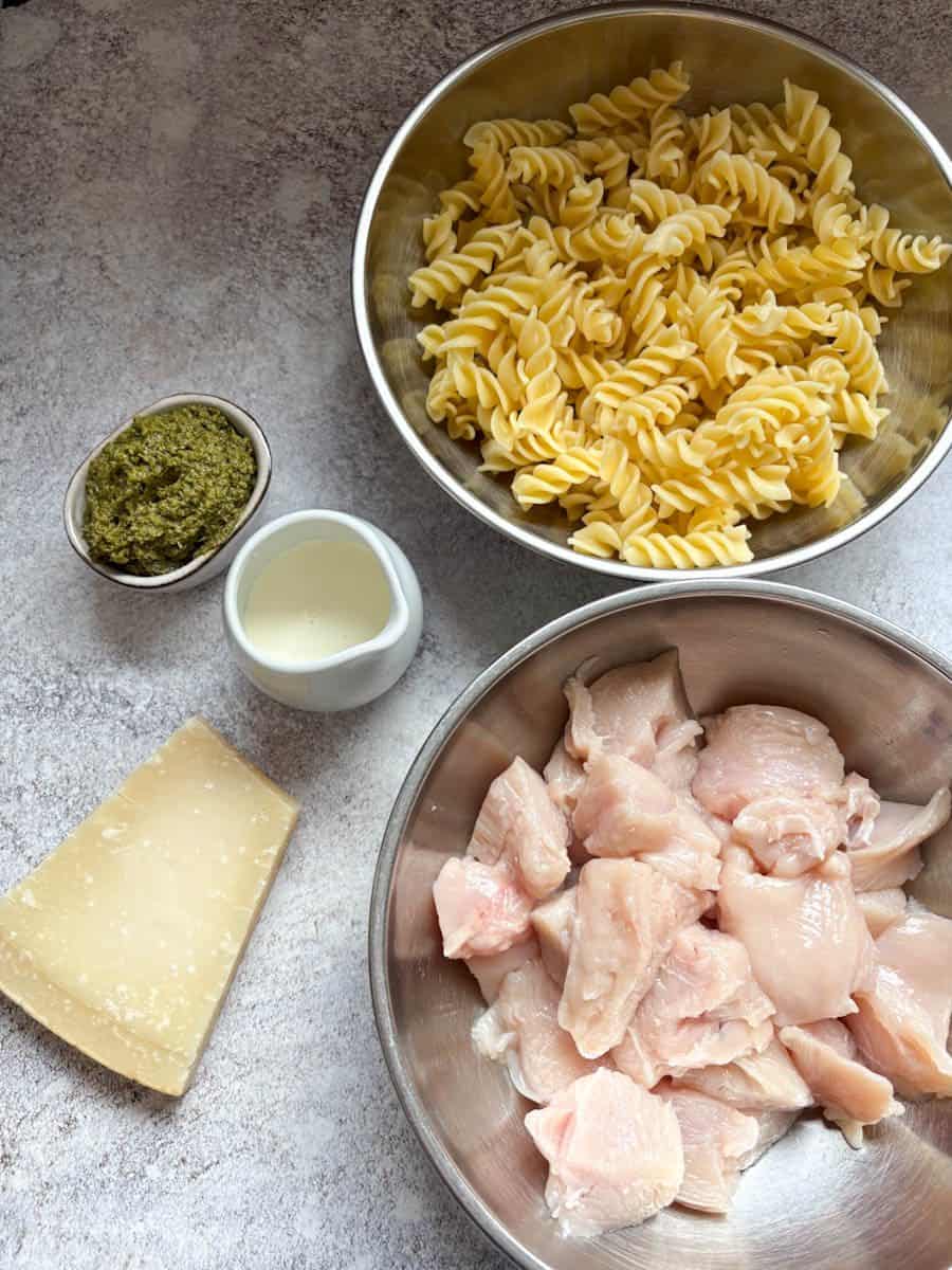 A silver bowl of raw chicken pieces, a silver bowl of Fusilli pasta, a pot of green pesto, a small white jug of cream and a piece of parmesan cheese on a white and grey mottled surface.
