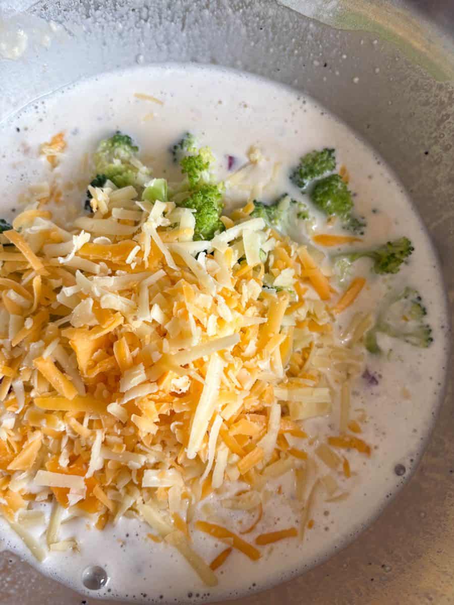 A silver bowl of eggs, milk, grated cheese, red onion and broccoli florets.