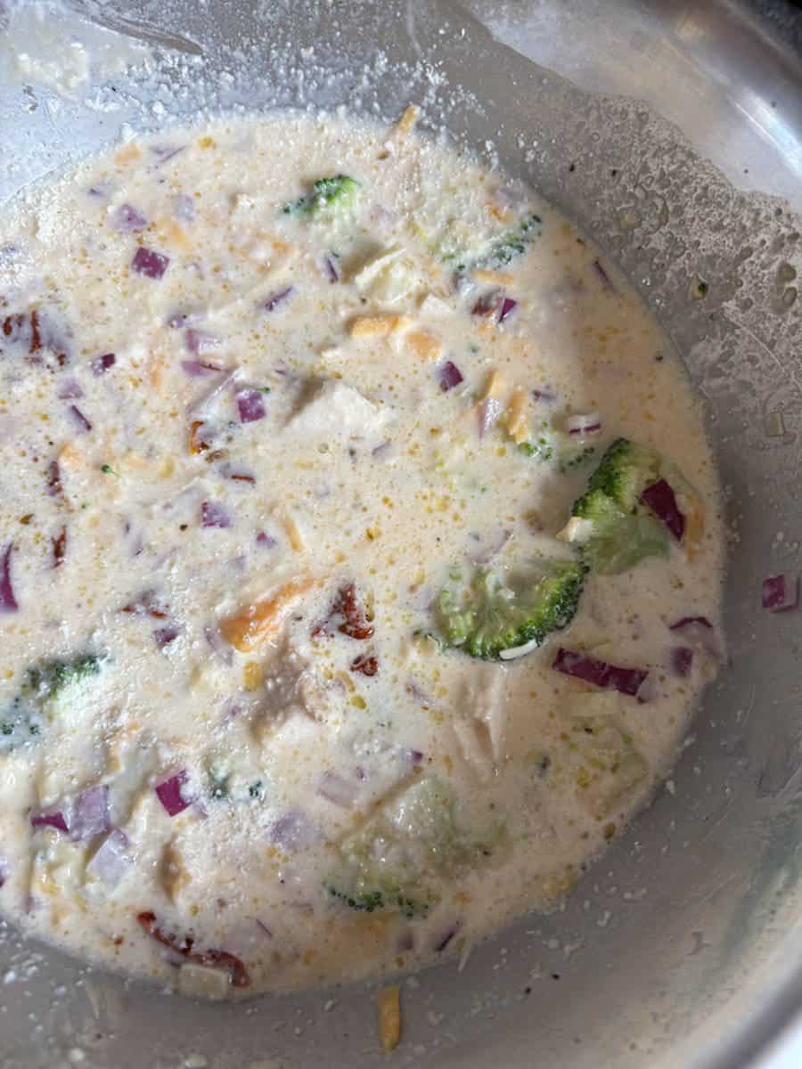 A silver bowl of whisked eggs, milk, broccoli florets, red onion and grated cheese.