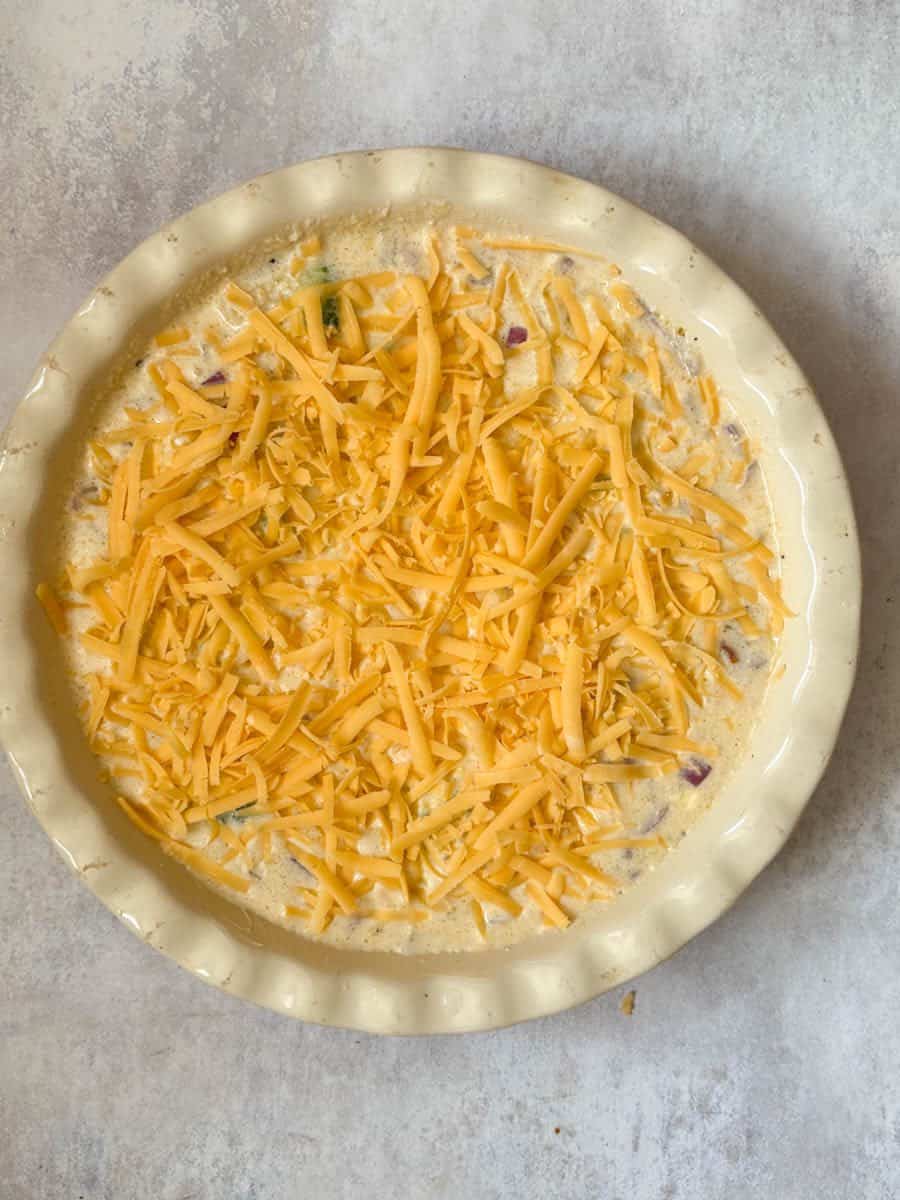An unbaked crustless quiche sprinkled with grated cheese in a white pie dish.