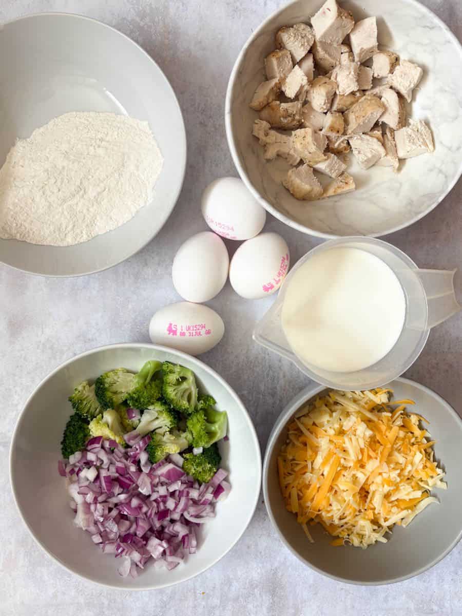 A selection of ingredients in bowls for making a crustless quiche - including flour, diced chicken, broccoli, red onion, grated cheese, a jug of milk and 4 eggs.