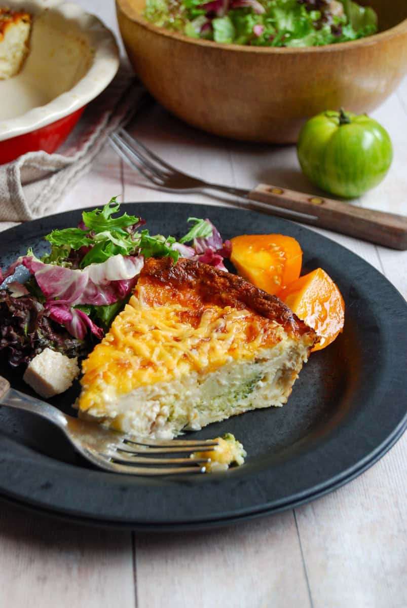 A slice of crustless quiche with a mixed salad and an orange sliced tomato on a black plate. A wooden bowl of salad, a green tomato and a fork with a wooden handle sits in the background.