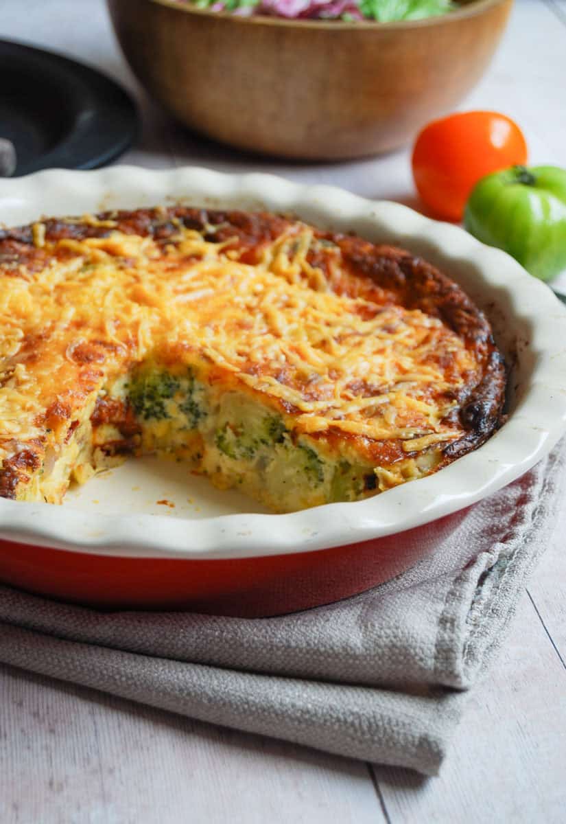 A crustless quiche filled with broccoli, sun dried tomatoes and chicken and topped with cheese on a red and white pie dish. A wooden bowl of salad (partially seen) and two tomatoes - one green and one orange - is behind the dish.