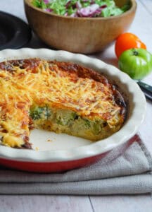 A crustless quiche filled with broccoli, chicken, sun dried tomatoes and topped with cheese in a white and red pie dish. A wooden bowl of salad and two tomatoes - one green and orange - sits behind the pie dish.