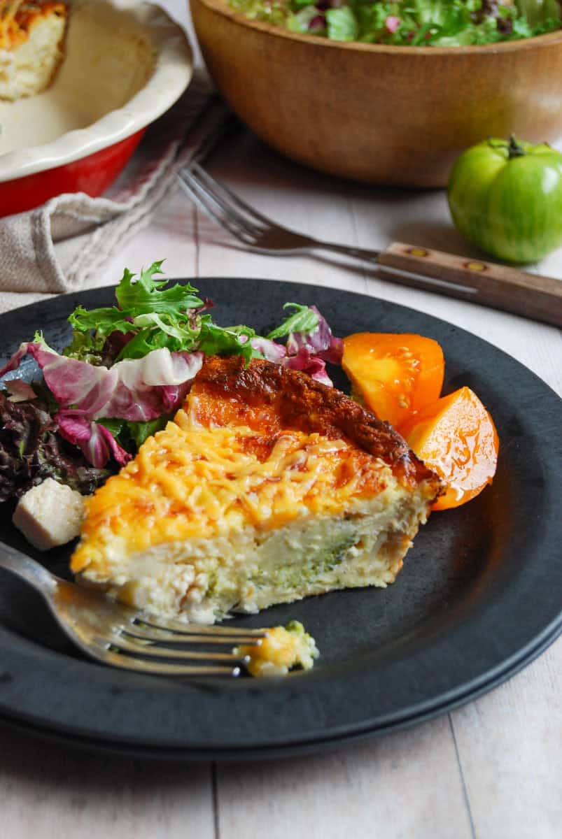 A slice of crustless quiche with a salad and a sliced orange tomato on a black plate with a silver fork. A wooden bowl of salad, a fork with a wooden handle and a green tomato sits in the background.