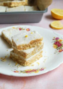 Three lemon and coconut bars stacked on top of each other on a white plate with a pink and yellow flower decoration. Lemon slices and a tin of the lemon bars can be partially seen in the background.