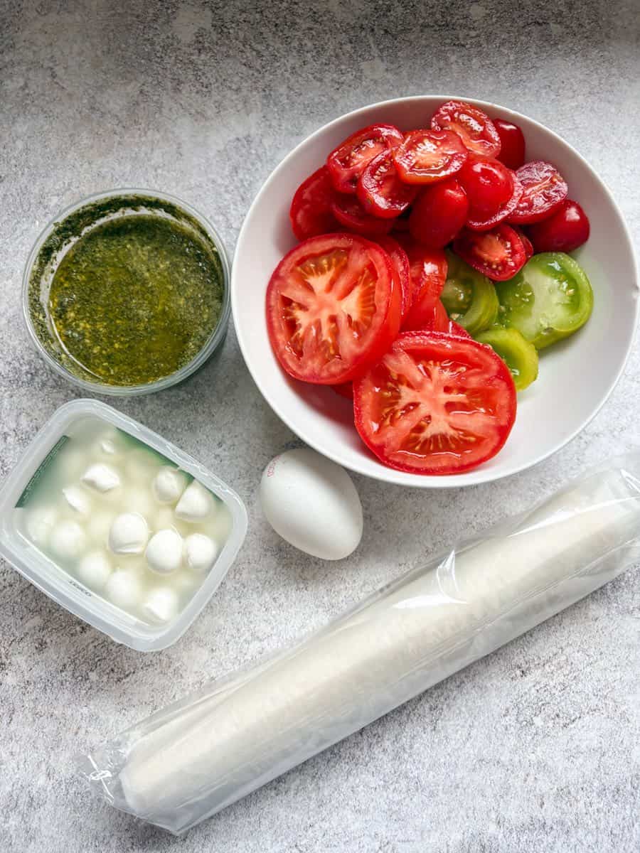 A white bowl of sliced red and green tomatoes, a pot of green pesto, a tub of mozzarella pearls, an egg and a packet of ready rolled puff pastry.