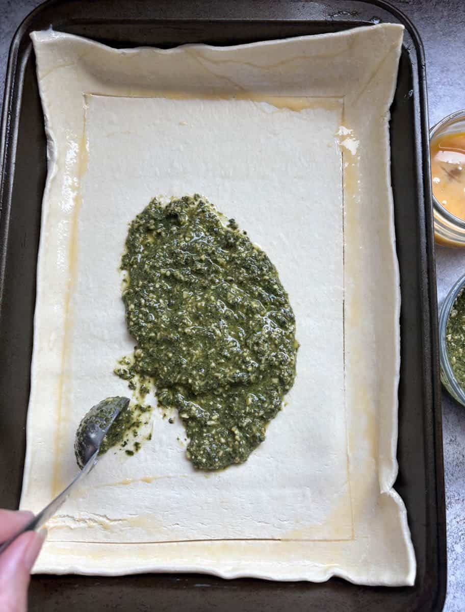 Green pesto being spread on a sheet of ready rolled puff pastry.