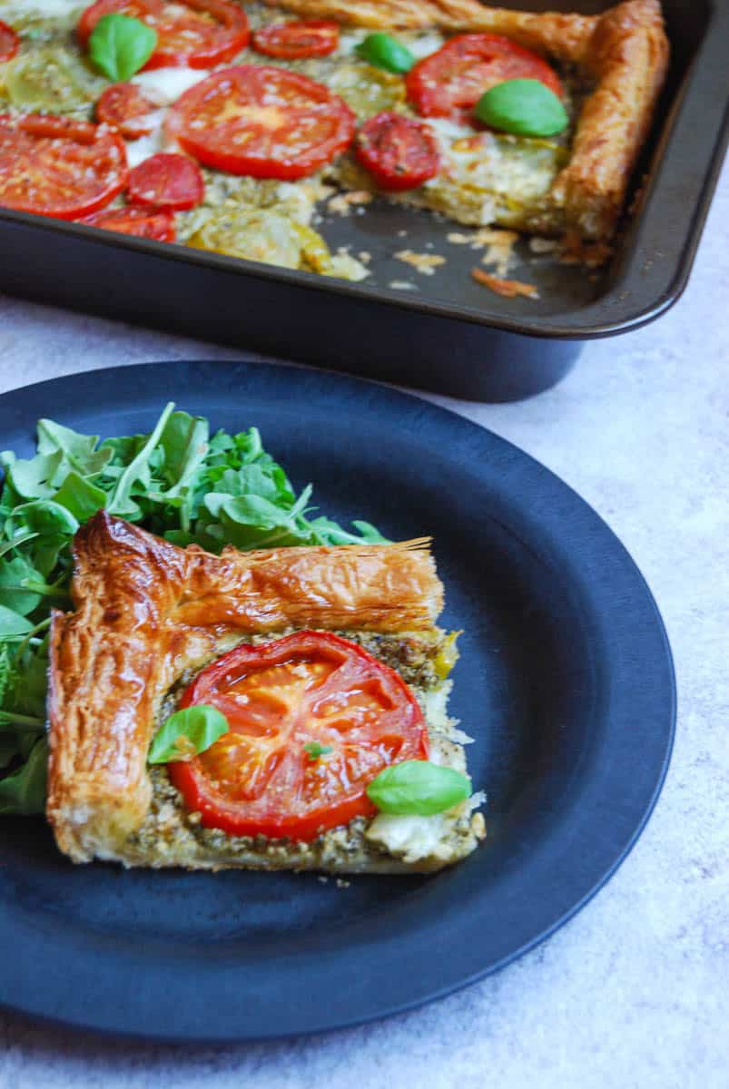 A slice of tomato pesto puff pastry tart on a black plate with fresh rocket leaves. A large tomato tart with a slice removed sits above the plate.