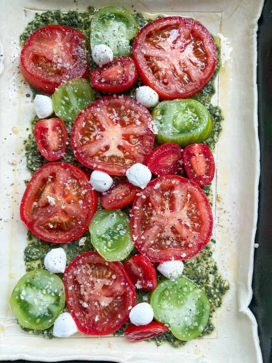 A sheet of puff pastry on a baking sheet spread with green pesto and topped with sliced red and green tomatoes and mozzarella pearls.