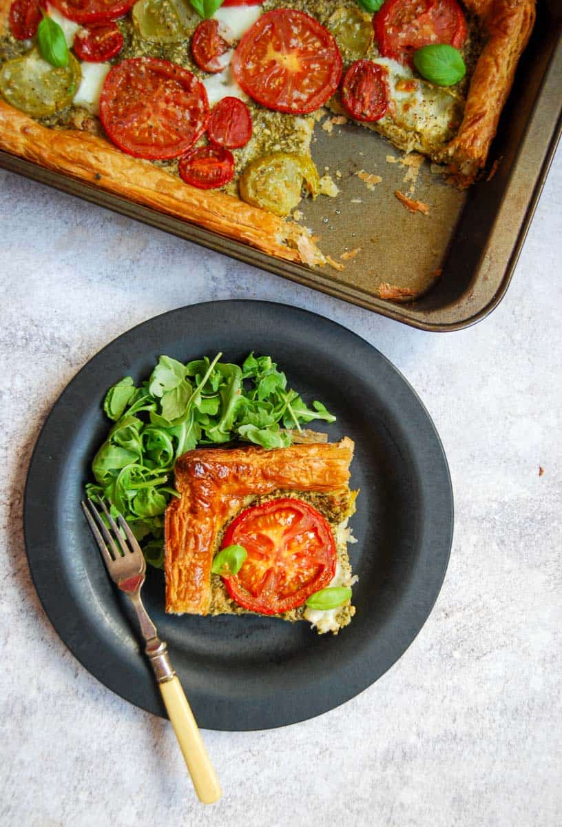 A slice of puff pastry tomato tart topped with fresh basil leaves on a black plate with fresh rocket leaves. A silver fork with a yellow handle rests on the plate. A large tomato pesto tart with a slice removed sits above the plate.