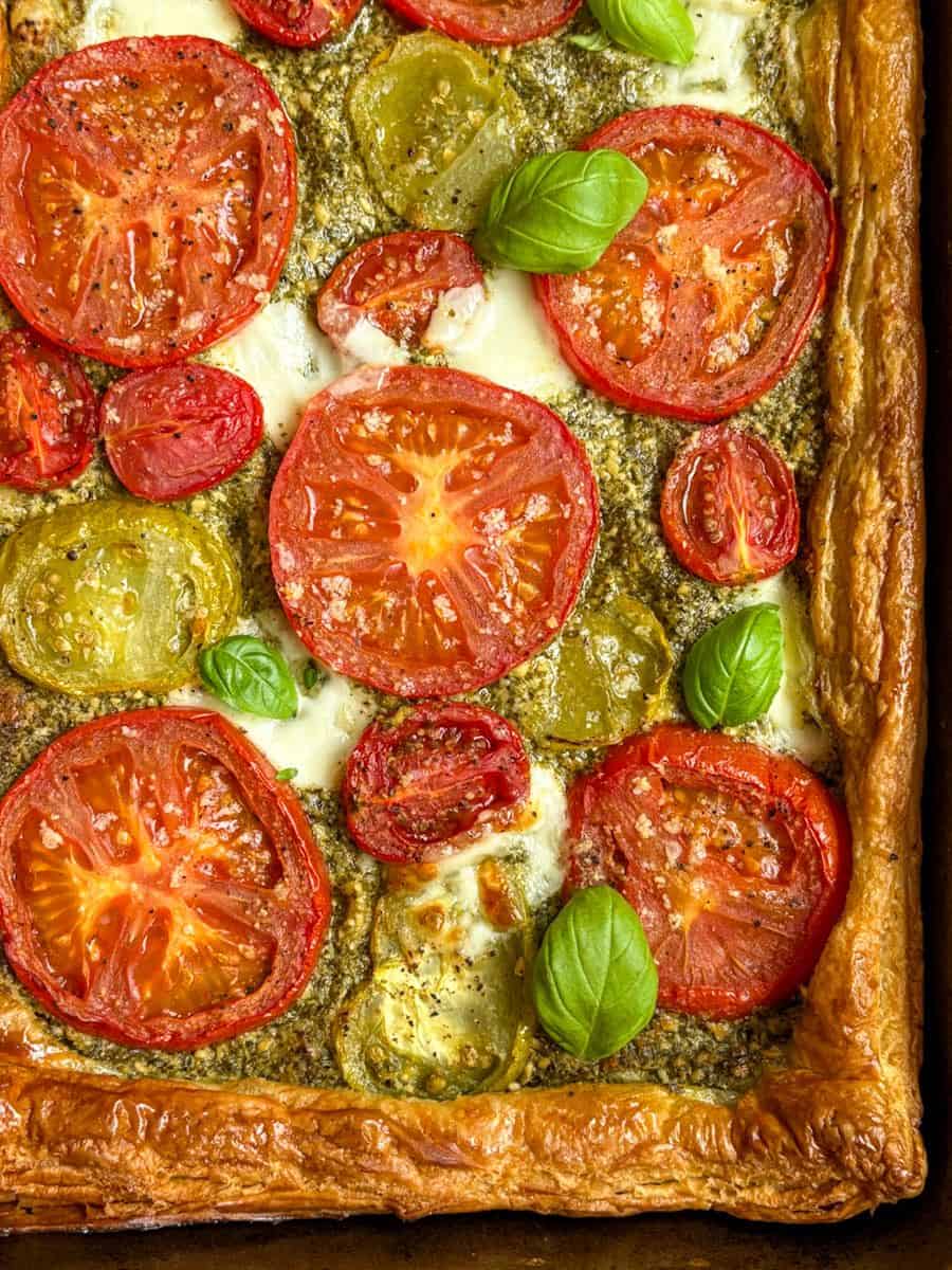 A close up photo of a puff pastry tomato, pesto and mozzarella tart strewn with fresh basil leaves.