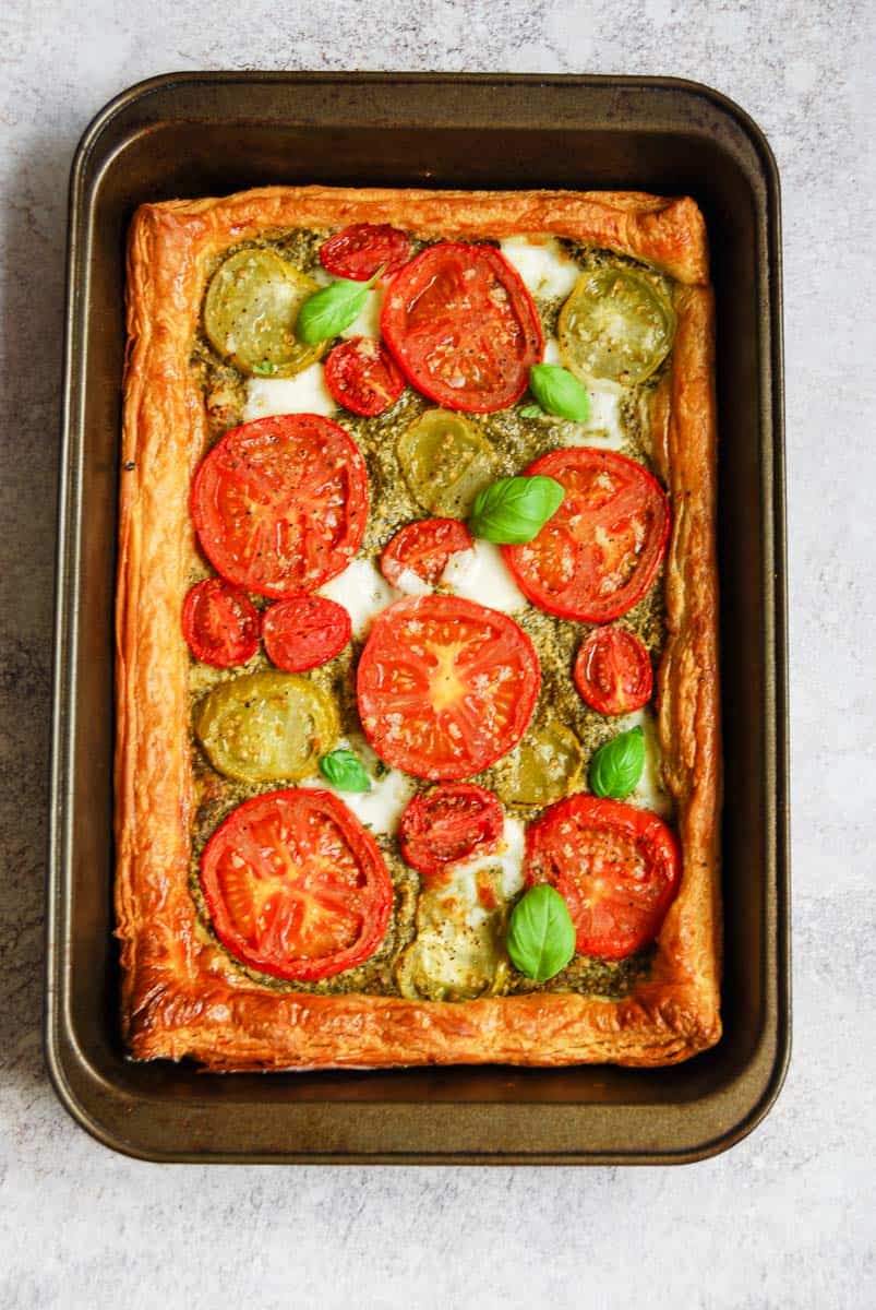 A fresh tomato and pesto puff pastry tart topped with fresh basil leaves in a rectangular baking tin.