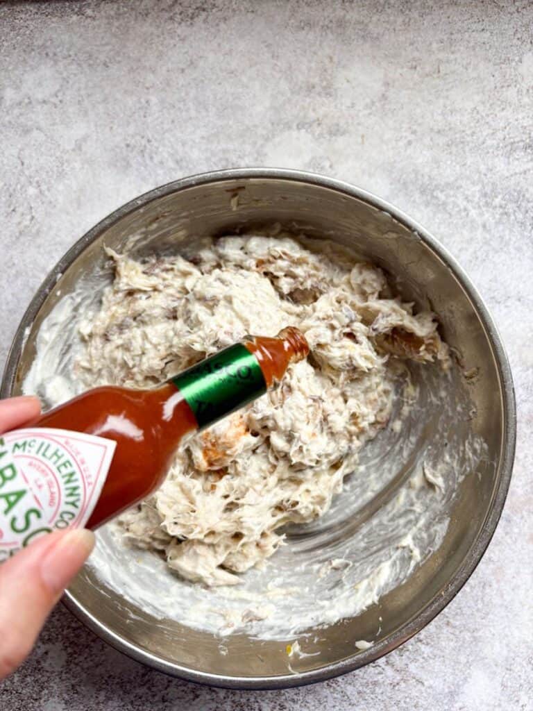 A hand holding a bottle of Tabasco sauce and a silver bowl of smoked mackerel fillets.