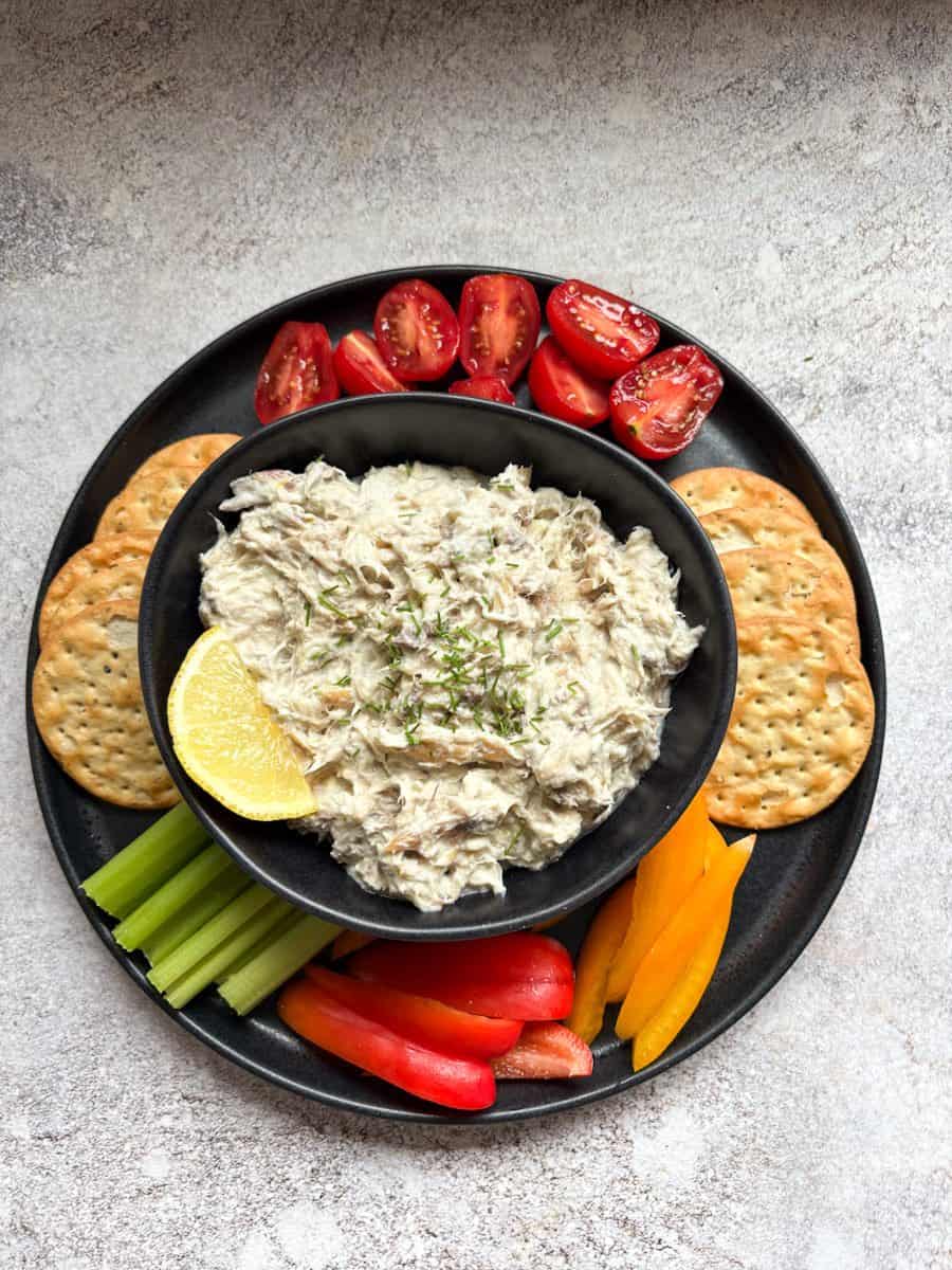 A black bowl of smoked mackerel pate sprinkled with chopped chives and a lemon slice and a black plate with cherry tomatoes, crackers, celery sticks and sliced red and yellow peppers.