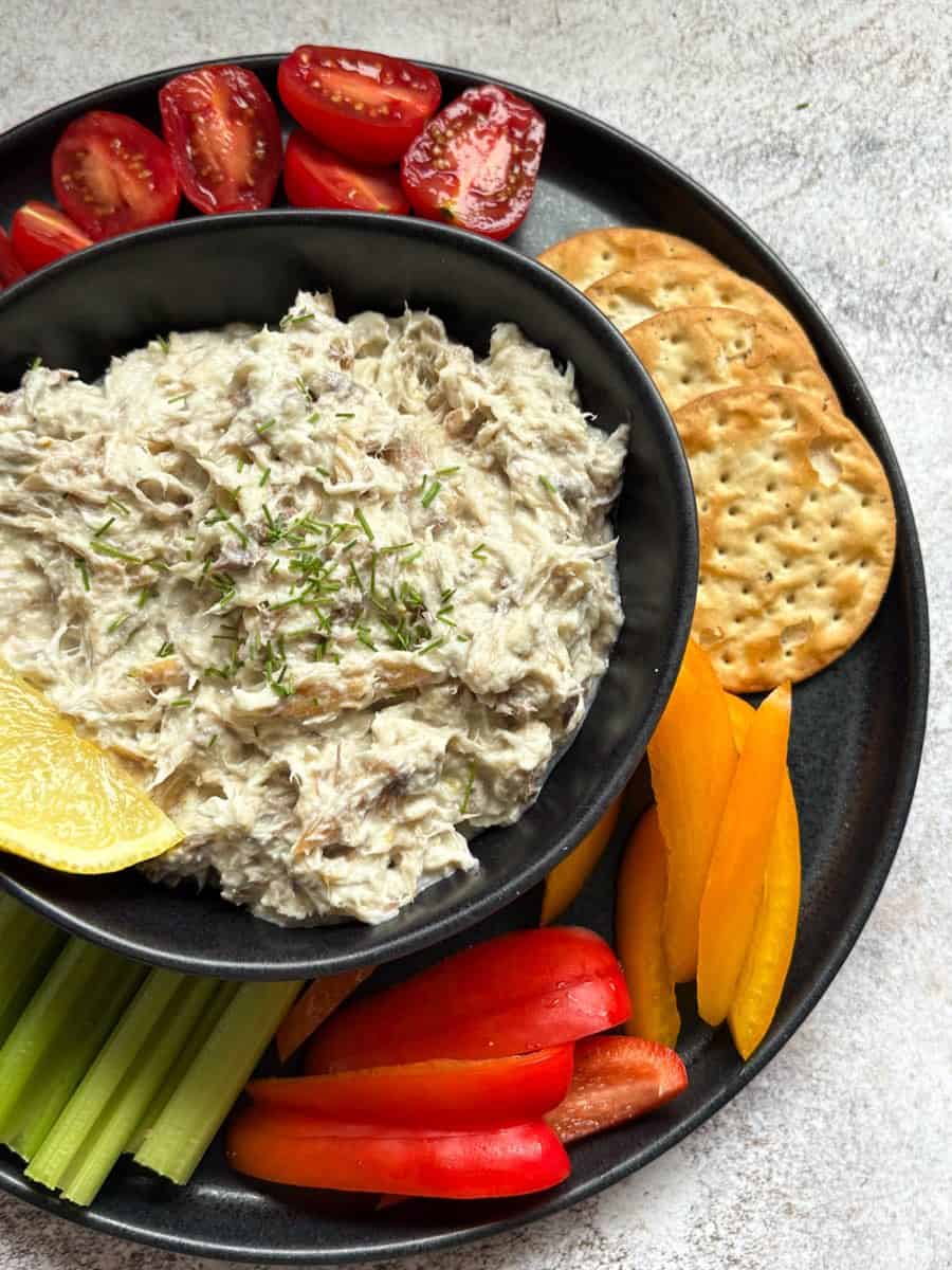 A black bowl of smoked mackerel pate sprinkled with chopped chives and a slice of lemon and a black plate of crackers, cherry tomatoes, celery sticks and sliced red and yellow peppers.