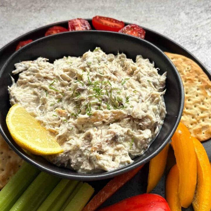 A black bowl of smoked mackerel pate sprinkled with chopped chives and a slice of lemon and a black plate of crackers, cherry tomatoes, celery sticks and sliced red and yellow peppers.