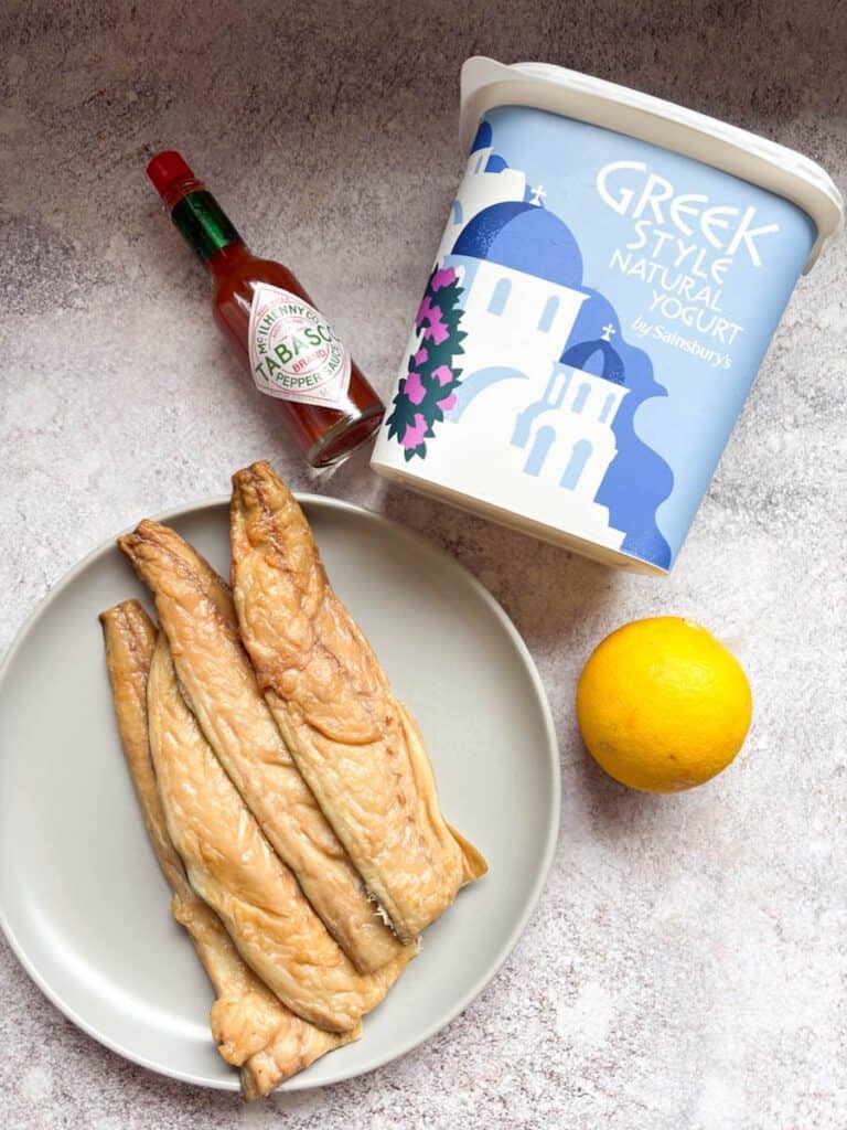 Four Mackerel fillets on a grey plate, a bottle of Tabasco sauce, a large tub of Greek yoghurt and a lemon on a grey surface.