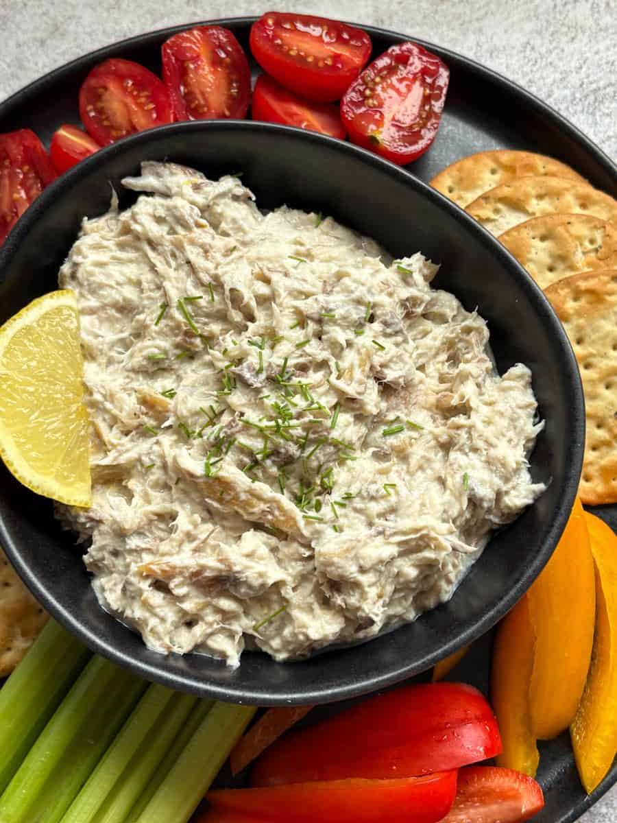 A black bowl of smoked mackerel pate sprinkled with chopped chives and a slice of lemon and a black plate of crackers, cherry tomatoes, celery sticks and sliced red and yellow peppers.
