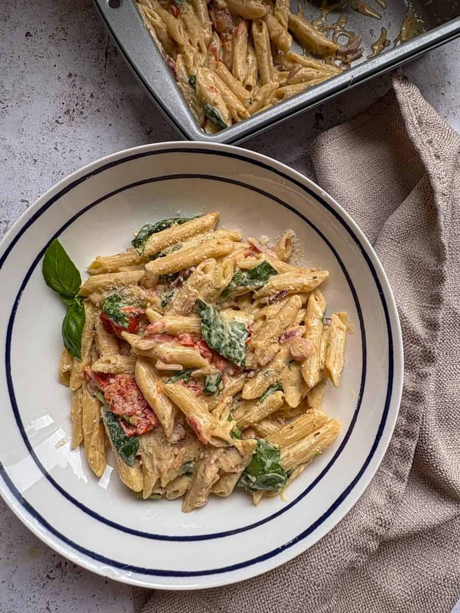 A white and blue bowl of penne pasta with Boursin cheese, cherry tomatoes, spinach and fresh basil and a beige linen napkin.