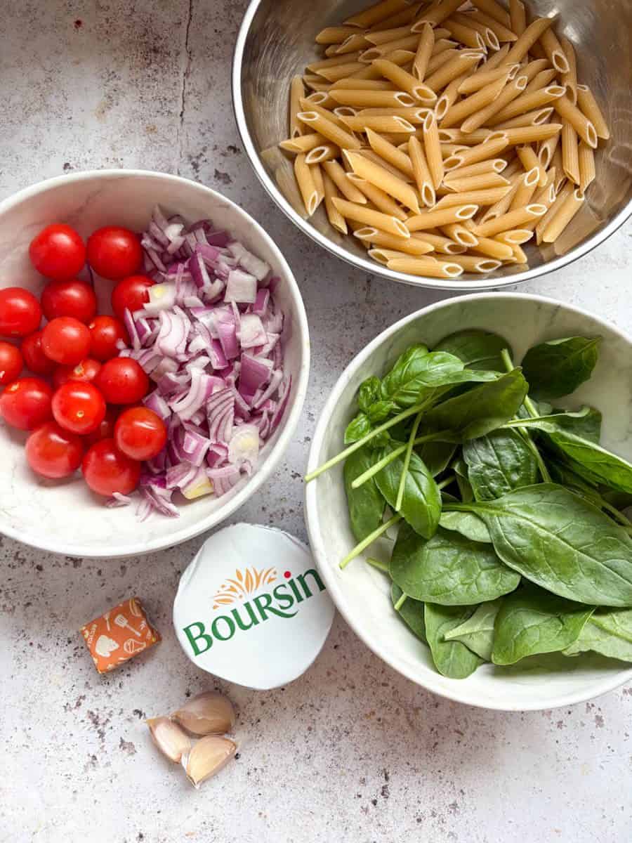 A silver bowl of Penne pasta, a white bowl of cherry tomatoes and chopped red onion, a white bowl of fresh spinach leaves, a packet of Boursin cheese, a chicken stock cube and three small cloves of garlic.
