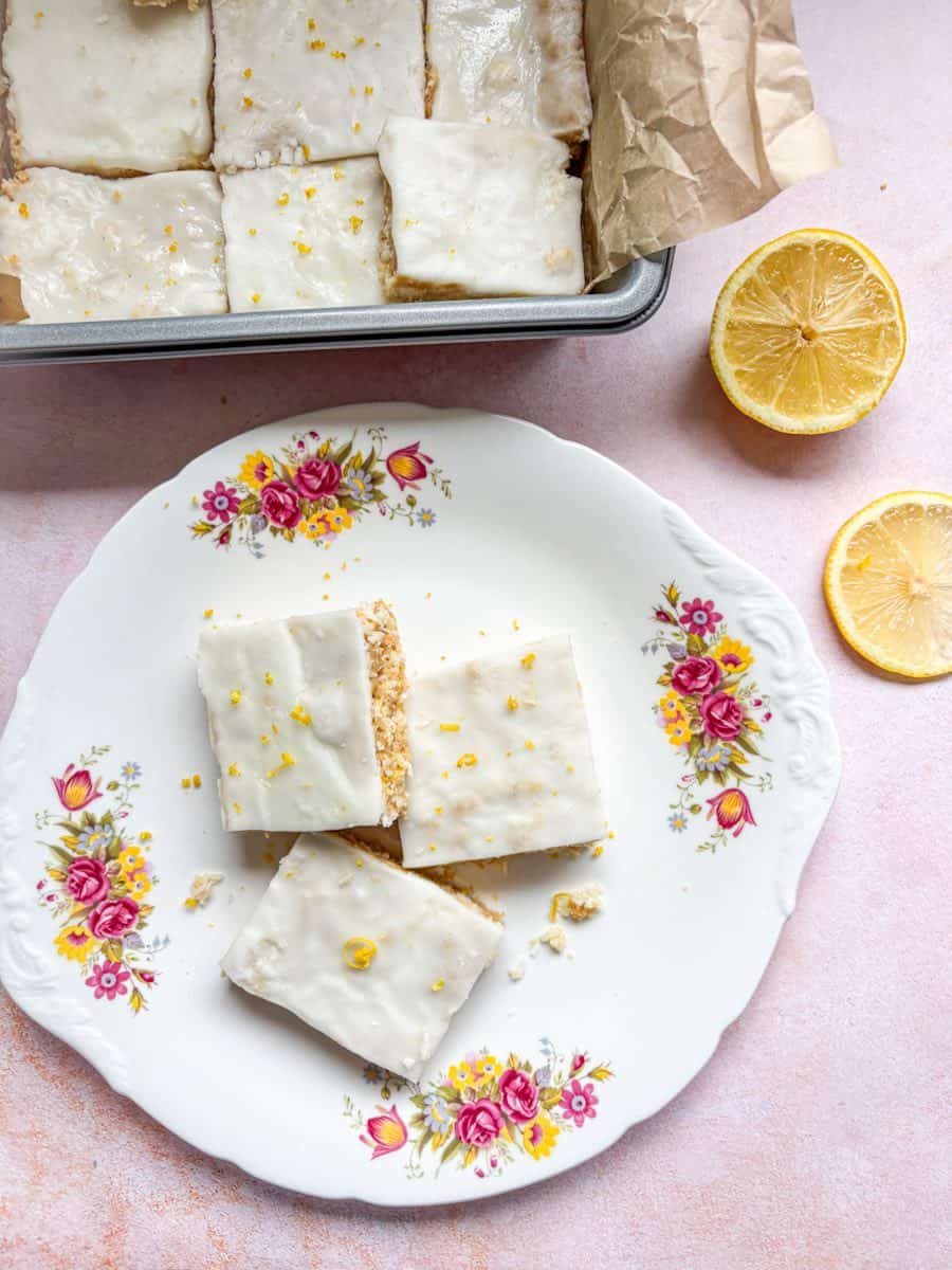 Three lemon and coconut bars on a white plate with a pink and yellow flower pattern. Lemon slices and a tray of the lemon bars sit above the plate.