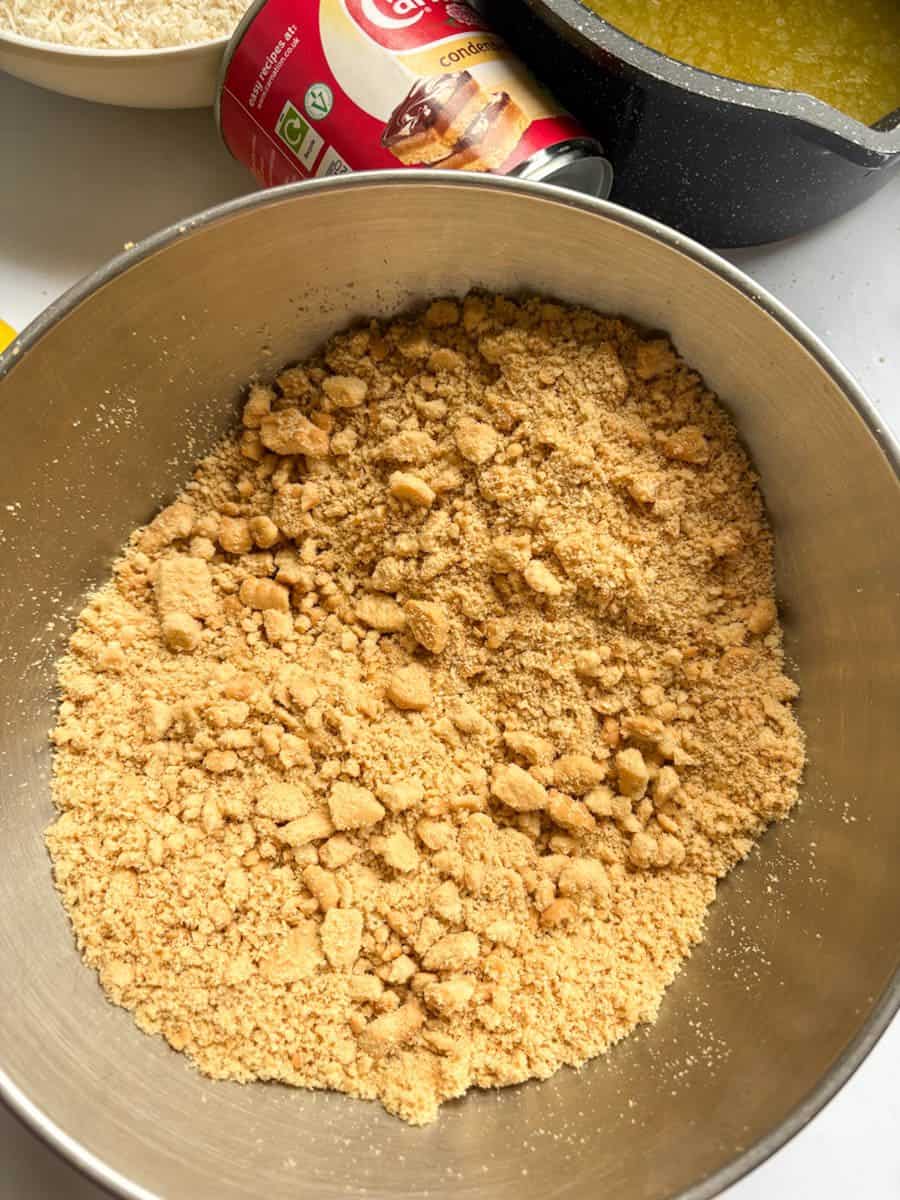 A silver bowl of crushed biscuits and a tin of condensed milk. A pan of melted butter can be partially seen.