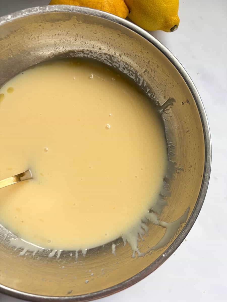 A silver bowl of condensed milk and melted butter with a gold spoon. Two lemons can be partially seen.